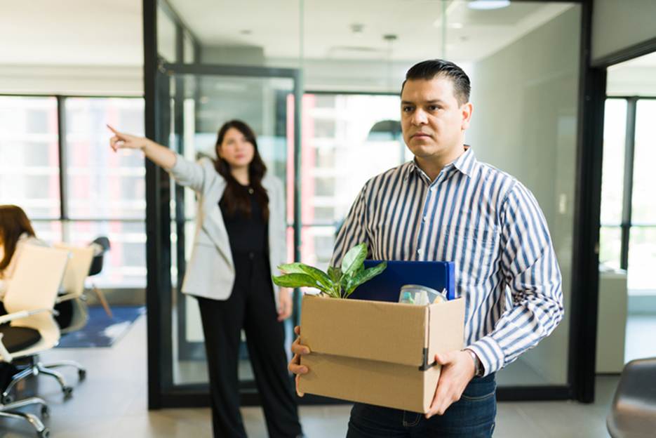 A man in an office holds a box of his belongings with a woman pointing away. The image is used as content for website.